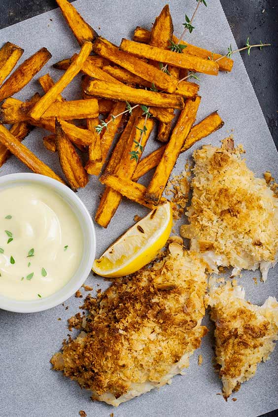 Coarsely crumbed fish fillets, a lemon wedge, sweet potato chips and a ramekin with white sauce on a speckled baking tray.