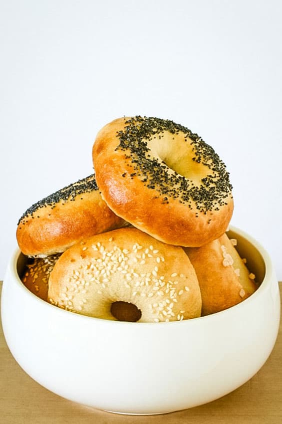 A white bowl with bagels, each topped with sesame seeds or poppy seeds or flaky salt, arranged on top of one another.