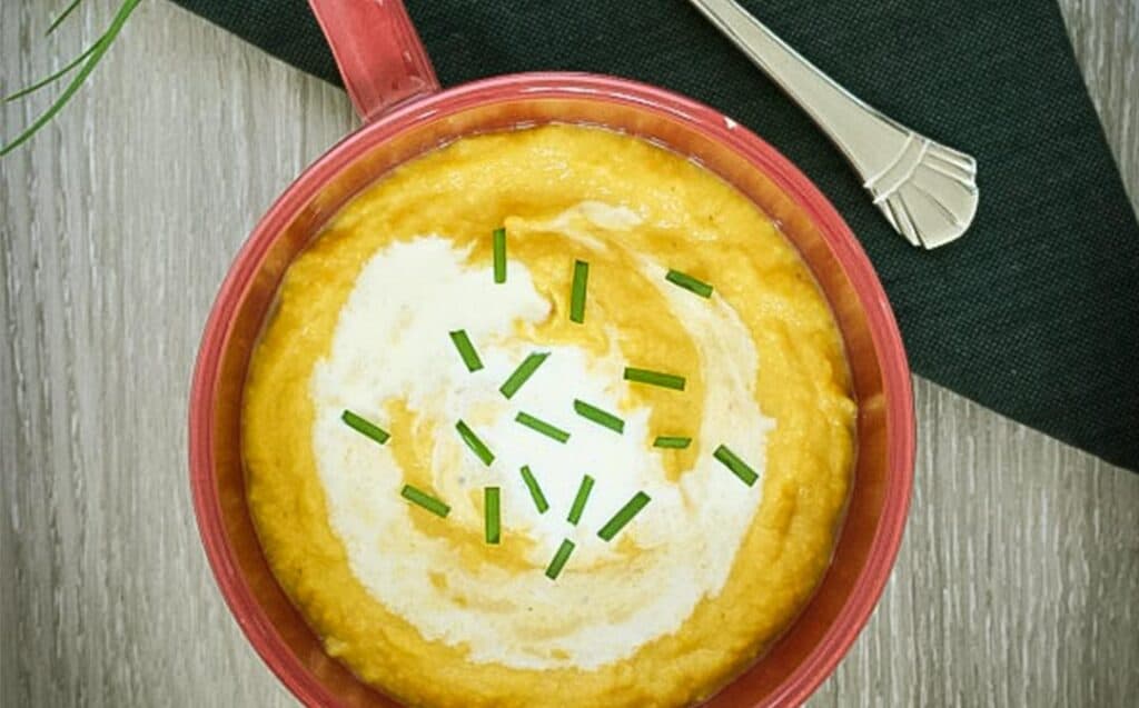 A red soup mug with thick, golden cauliflower and pumpkin soup garnished with chives