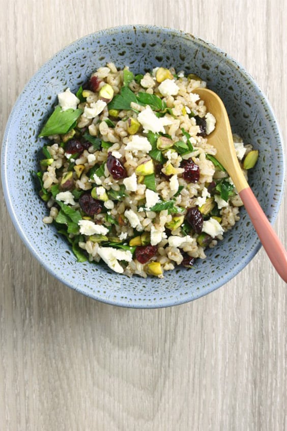 A speckled blue bowl on a pale wooden board, with a rice salad with cranberries, pistachios, feta and fresh herbs and a salad server.