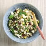 A speckled blue bowl on a pale wooden board, with a rice salad with cranberries, pistachios, feta and fresh herbs.