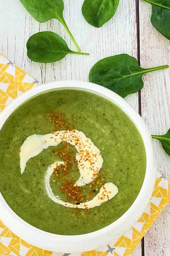 A bowl of green soup with a swirl of cream and a sprinkle of seasoning on a pale wooden surface