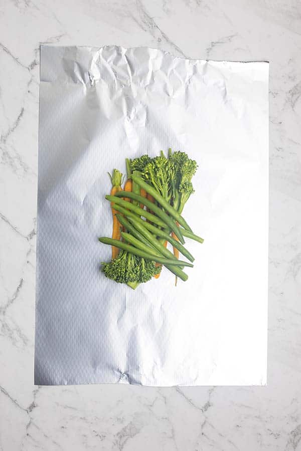 A rectangular piece of foil laid on a marble countertop with carrots, beans and baby broccoli