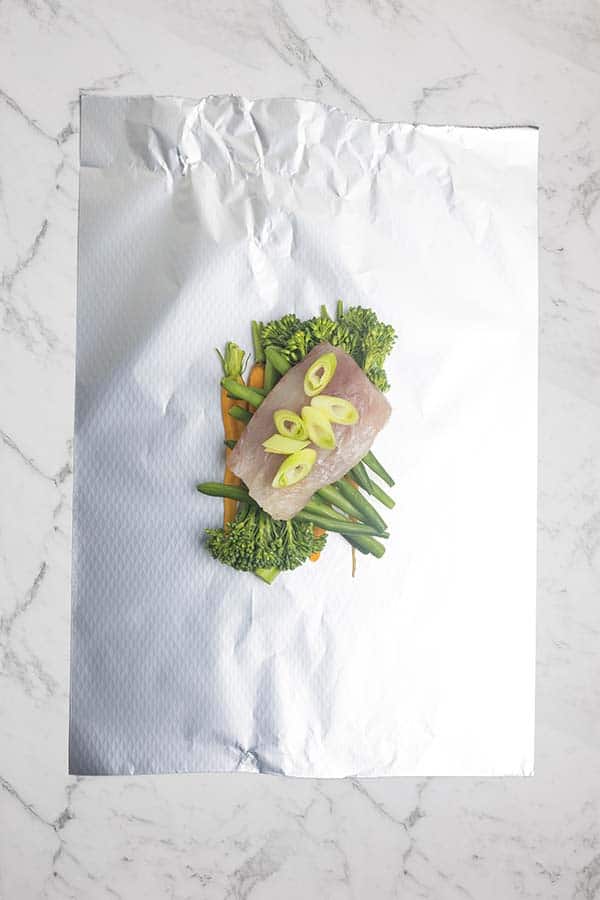 A rectangular piece of foil laid on a marble countertop with a piece of fish on a bed of vegetables topped with scallions