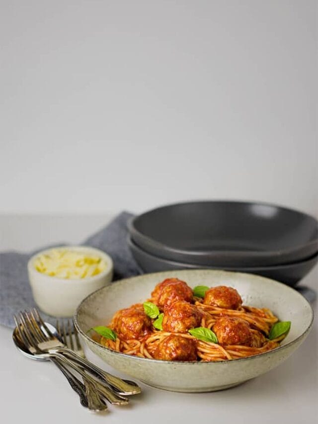 A picture of a bowl of spaghetti and meatballs in the foreground and bowls and grated cheese in a ramekin
