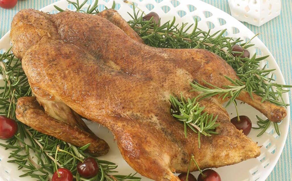 A white platter with cutout edges on a striped cloth with a whole roast duck, rosemary sprigs and cherries