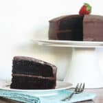 a wedge of frosted chocolate layer cake on a white plate in front of a cake stand with the rest of the cake