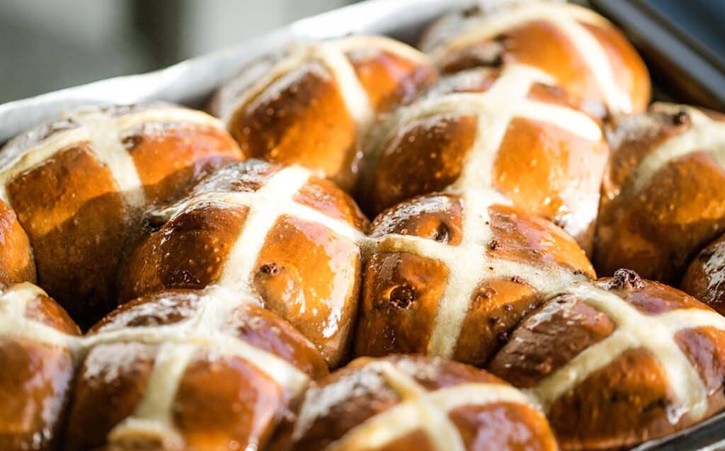 a close up image of golden brown glazed hot cross buns in a baking tin