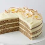 a close up image of a triple layered cake, with a quarter cut out to show the inside, on a white cake stand with thick, pale yellow frosting decorative apple slices on one side of the cake