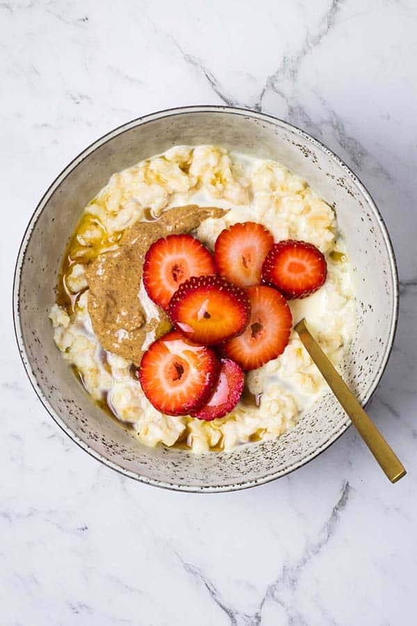 a bowl of cooked oatmeal with sliced strawberries, almond butter, maple syrup and a dash of cream