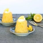 two individual steamed lemon puddings garnished with cream and lemon with dessert fork and a halved lemon on a dark linen covered counter