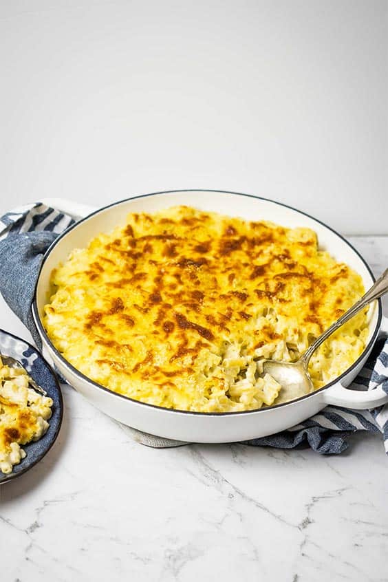 a round cream baking dish sitting on a blue kitchen cloth; baked macaroni and cheese is in the dish with a spoonful removed