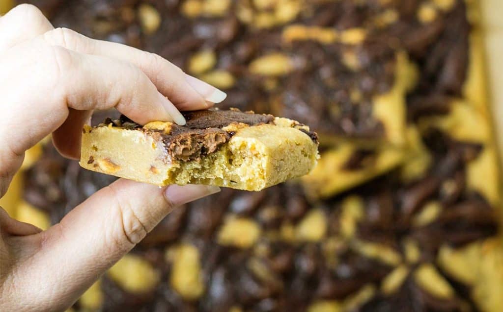 a close up image of a Nutella blondie being held between a person's fingers; a bit has been taken from the blondie