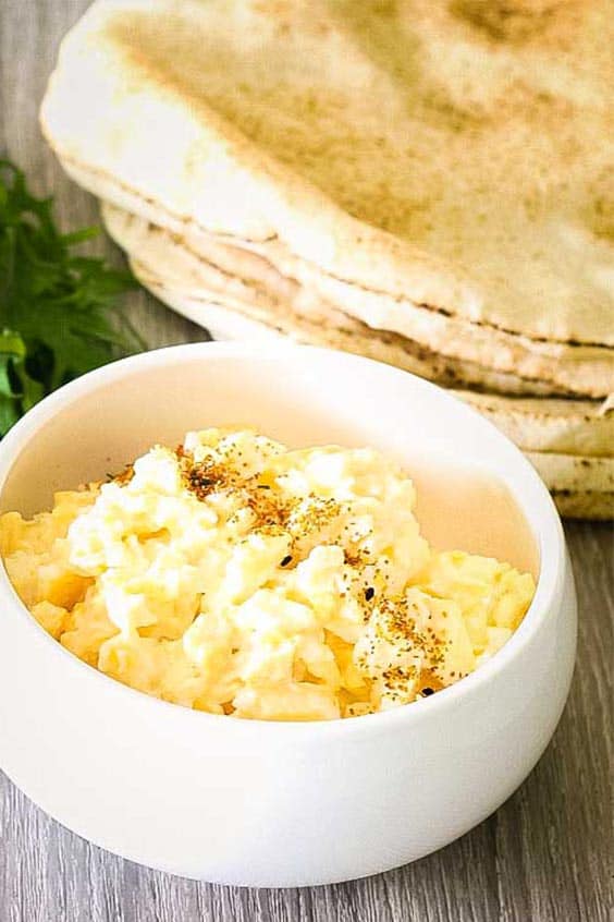 a white bowl full of scrambled eggs with a stack of flat breads in the background