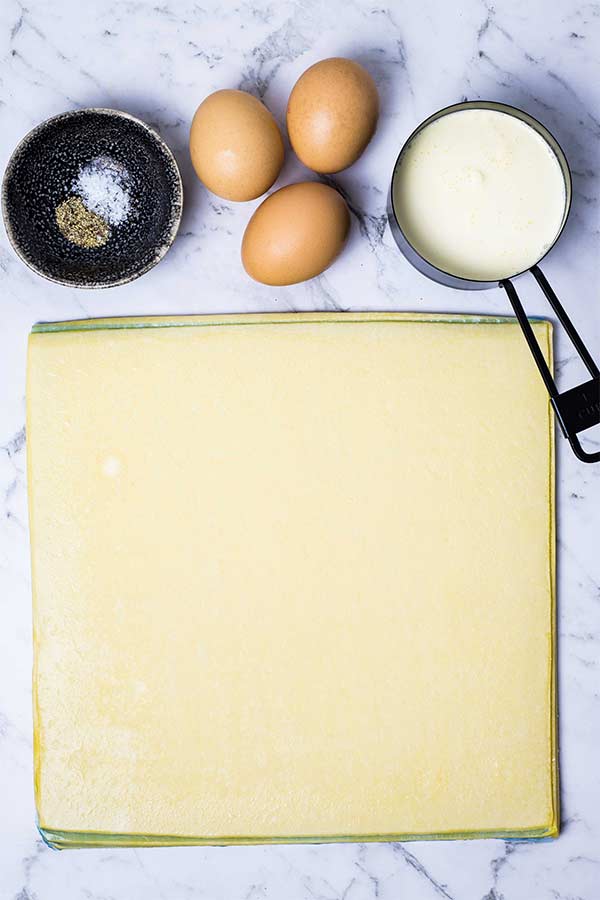 Sheets of shortcrust pastry, eggs, seasonings in a small dark bowl and a measuring cup with cream