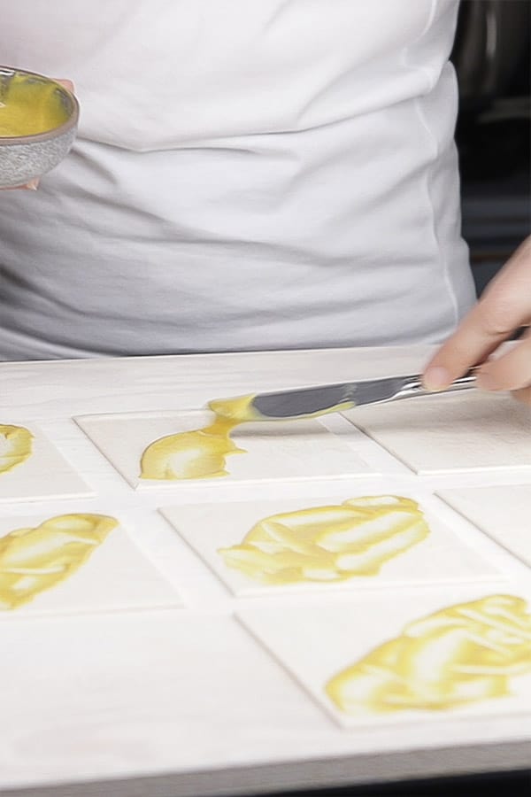 a yellow sauce being spread diagonally on the squares of pastry