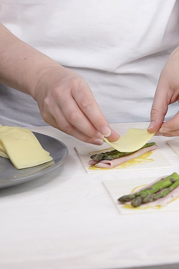 hands putting a rectangle of cheese on top of asparagus and ham on pastry