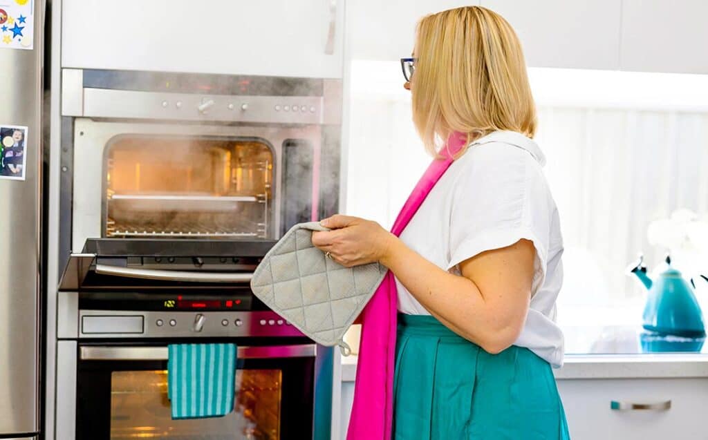 A woman with blonde hair, wearing a white shirt and green skirt, stands in front of an open steam oven. She holds a pot mitt, ready to remove a hot pan.