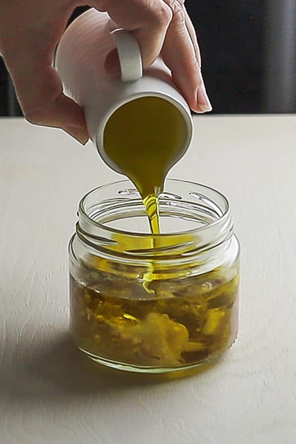 Oil being poured out of a small jug into a glass jar with dressing ingredients