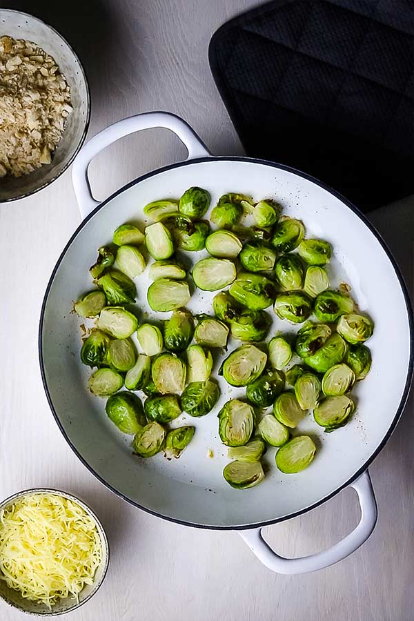 Halved brussels sprouts in a white enamel pan with two handles. Next to the sprouts is some shredded cheese and breadcrumbs, each in a small ceramic dish.