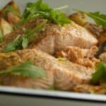 A close up view of the texture of roasted salmon with lemon and burnt butter in a baking dish along with rocket and roast potatoes