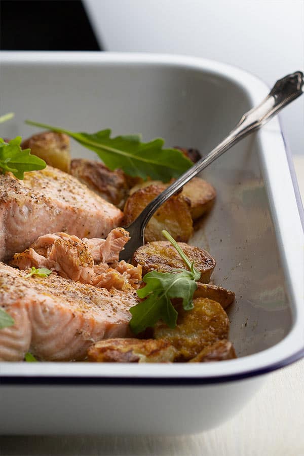 An enamel roasting dish with roasted halved potatoes, rocket, and a silver fork in a fillet of roasted salmon with lemon and burnt butter