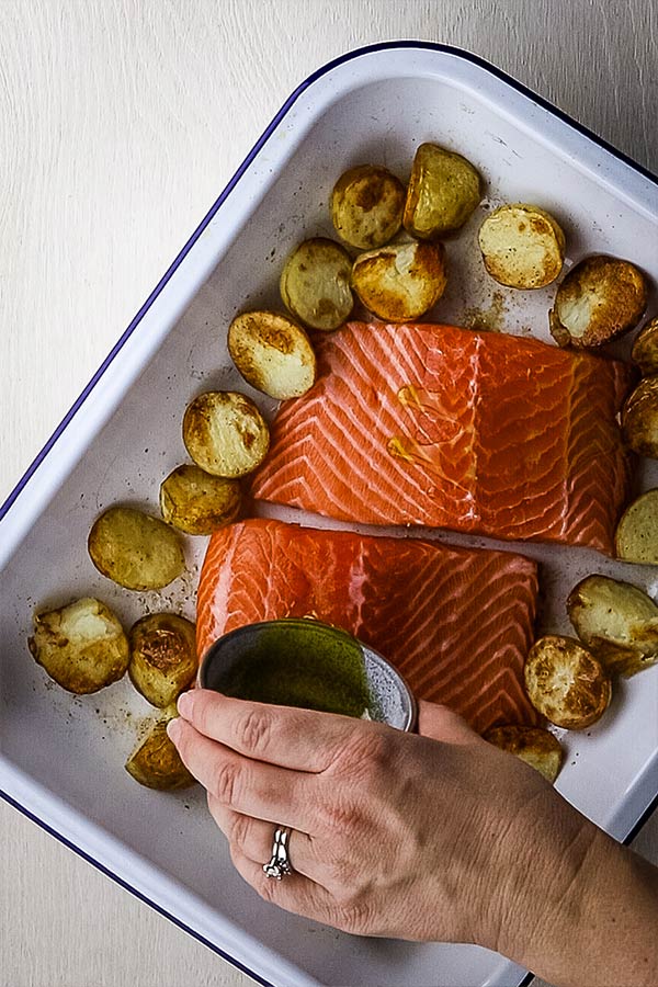 Lemon and burnt butter being poured over thick salmon fillets and partially roasted potatoes in a roasting dish