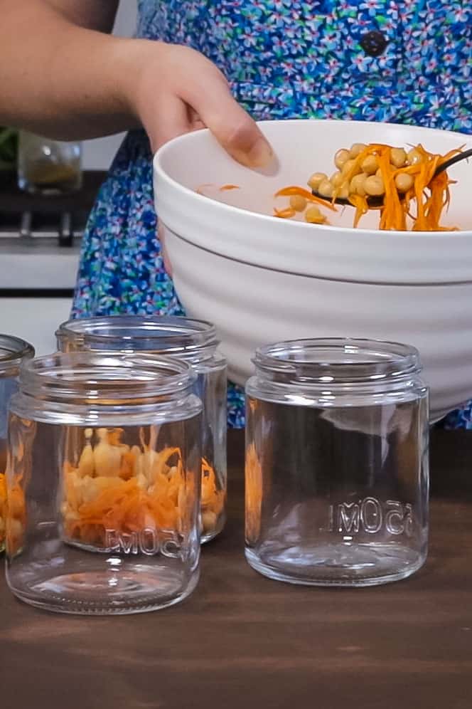 a woman's hand holds a large white mixing bowl filled with dressed chickpeas. The chickpeas are being spooned into large glass jars to make layered salad.