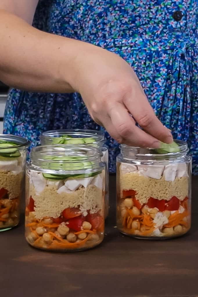 a woman's hand lays slices of cucumber into a jar full of layered chickpea and couscous salad