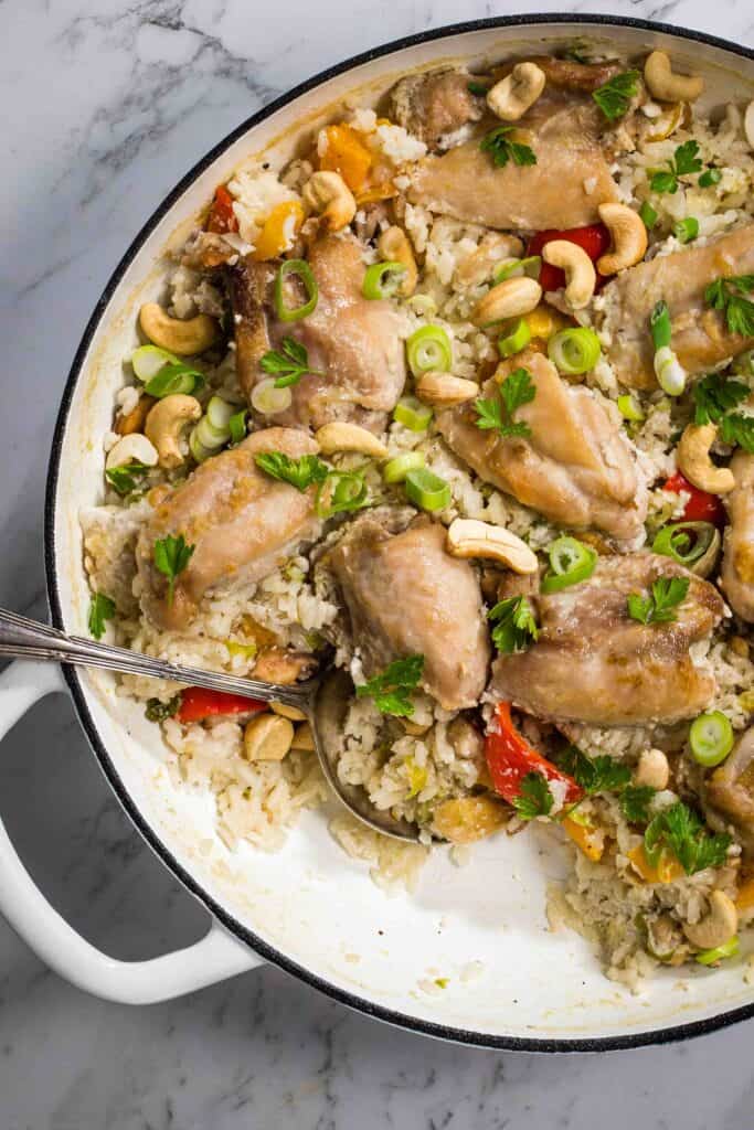 A white cast iron casserole dish filled with coconut chicken and rice, garnished with scallions. Pieces of chicken and bells peppers are visible on the top of the rice.