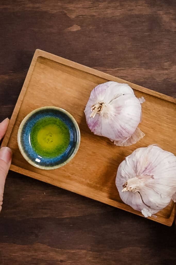 two heads of garlic sit next to a small blue dish filled with olive oil, on a wooden tray.