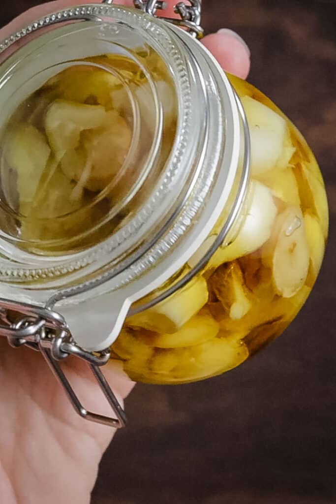 a swing top jar with white rubber seal, filled with roasted garlic cloves and olive oil. The jar is being held up to the camera with a hand visible behind it. 