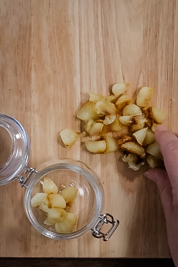 roasted garlic cloves being squeezed out of the skins, onto a wooden chopping board. A small glass jar with a few cloves of roasted garlic sits on the board.