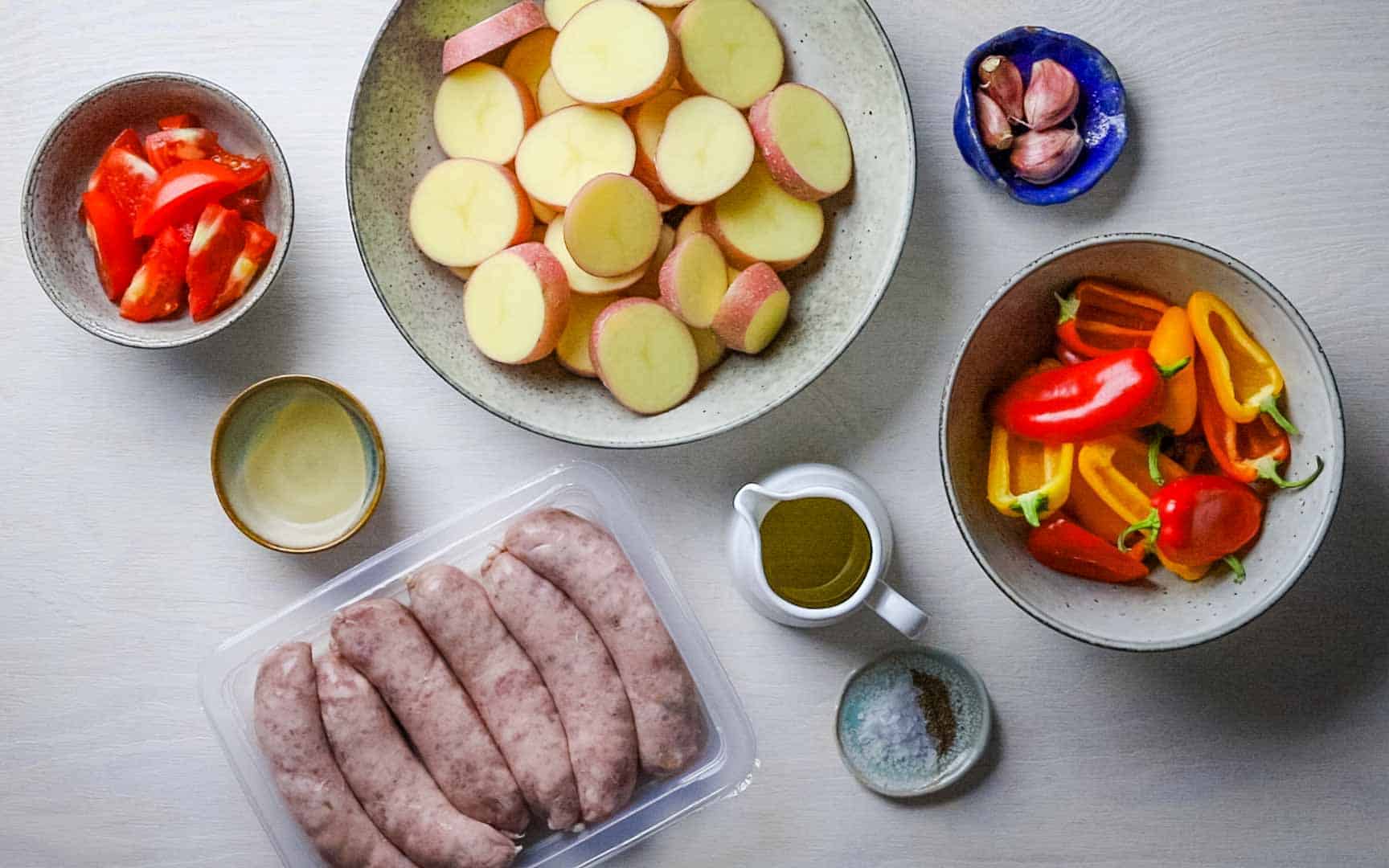 Bowls of chopped tomatoes, potatoes, peppers and garlic on a white wooden countertop, with a tray of raw sausages alongside the bowls.