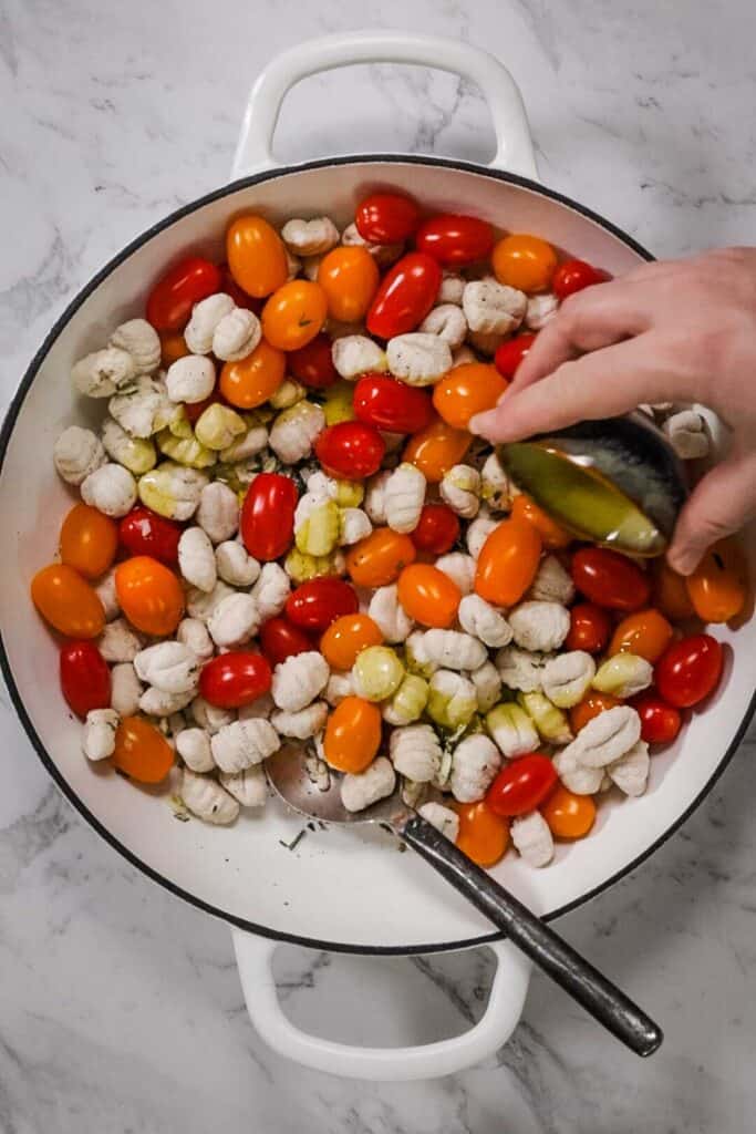 A large white cast iron baking dish with two handles; in the dish is frozen gnocchi, ripe grape tomatoes, garlic and rosemary. A hand holds a small blue dish of olive oil, pouring it over the top of the other ingredients. 