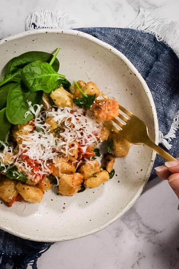 A blue woven napkin sits on a marble counter. Atop the napkin is a white dish with baked vegetarian gnocchi, parmesan cheese and red pepper flakes on top. A gold fork is holding two pieces of gnocchi next to the dish.