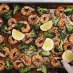 A sheet pan full of roasted broccoli and shrimp in soy ginger sauce. A hand places fresh lemon wedges around the pan for serving.