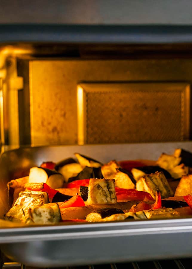 The inside of a stainless steel oven, where a warm yellow light casts a glow onto a tray full of roasted summer vegetables.