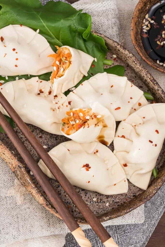 6 steamed chicken dumplings arranged on a brown ceramic plate, with steamed leafy greens and soy dipping sauce set to the side.