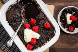 A rectangular, white enamel baking dish is filled with freshly baked steam oven chocolate pudding and garnished with thick cream and fresh raspberries. One serve has been scooped out of the dish and is in a round white bowl alongside.