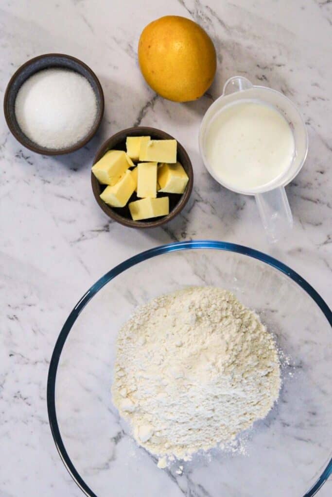 How to make lemon scones; a marble countertop with bowls of flour, butter, sugar and buttermilk, and a whole lemon