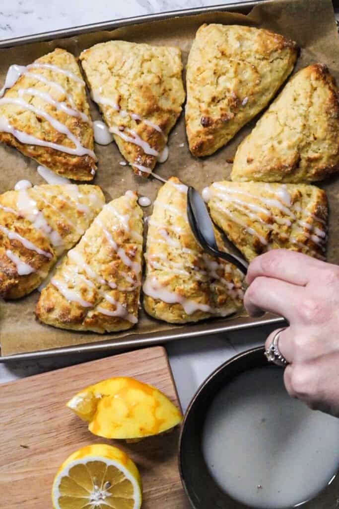A hand holds a black dessertspoon, drizzling lemon glaze over the tops of freshly baked lemon scone triangles.