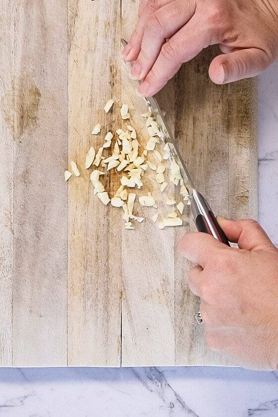 chopping cloves of garlic using knife atop a wooden chopping board