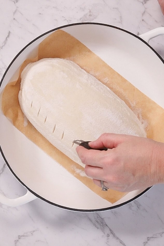 a hand holds a bread scoring razor, making small cuts into an unbaked loaf of steam oven sourdough bread