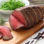 roast beef on top of a wooden cutting board with a bowl of lettuce on side