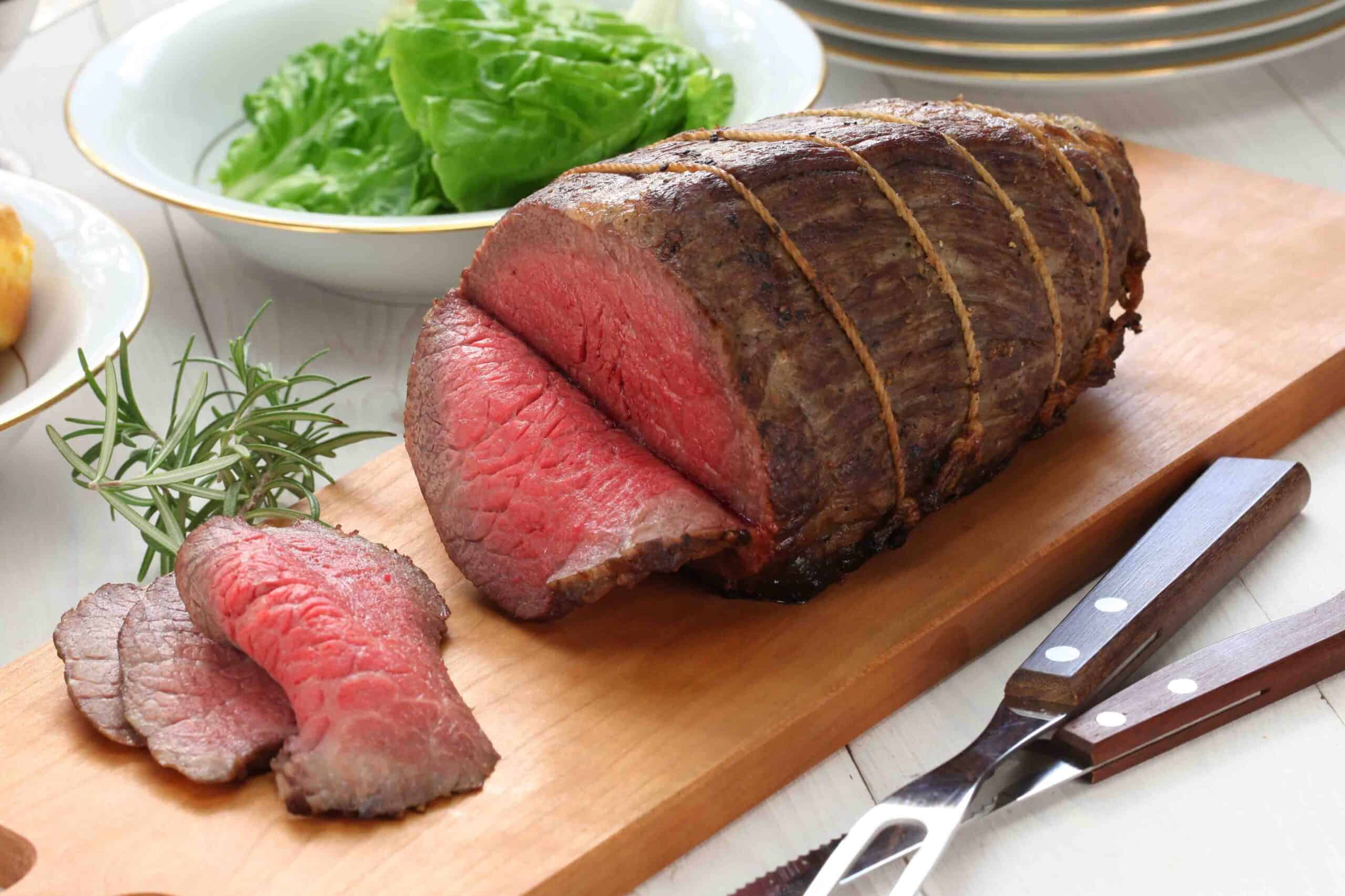 roast beef on top of a wooden cutting board with a bowl of lettuce on side