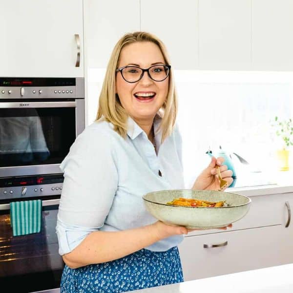 Emily stands in front of a wall mounted steam oven. She is wearing a blue top and skirt, and is holding a large bowl of steam oven roasted vegetables and a fork. Behind her is a white kitchen countertop and cabinets.
