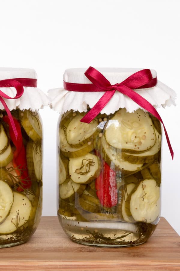 A tall glass jar with sliced pickled cucumbers and peppers. The jar has a white fabric cover and a red ribbon tied around the top. 