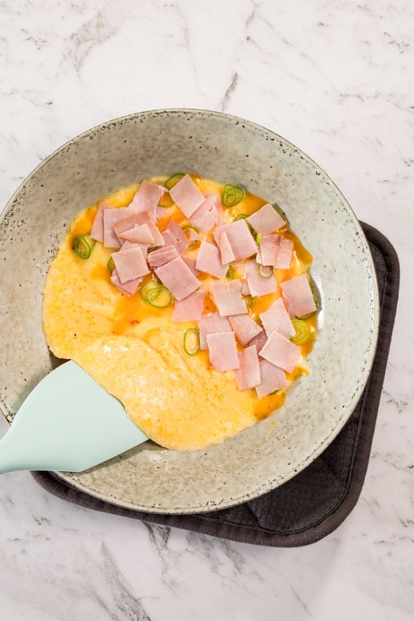 A grey ceramic bowl viewed from above, with a steamed omelette inside it. The omelette is topped with melted cheese, ham and green onions, and a blue spatula can be seen lifting it up to fold over on itself. 