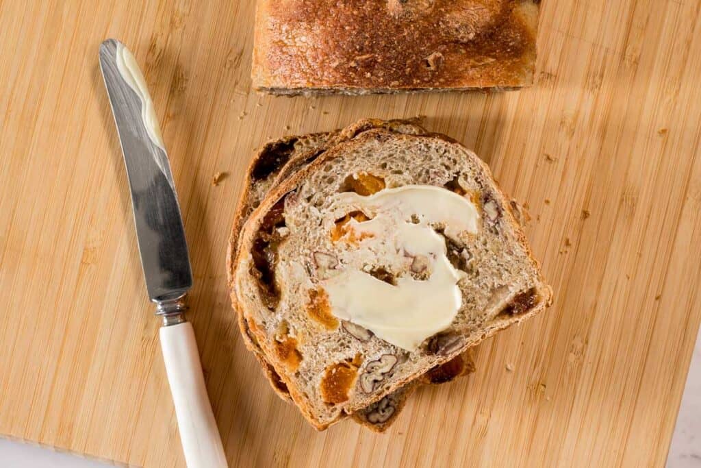 a slice of fruit and nut bread that's been baked in a steam oven, with a smear of butter across the surface and the butter knife alongside